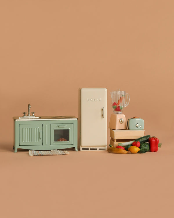 The Maileg Kitchen Set features a green sink and oven, cream refrigerator, blender, toaster, vegetables, fruit, and a striped rug—miniature accessories arranged on a simple beige background.