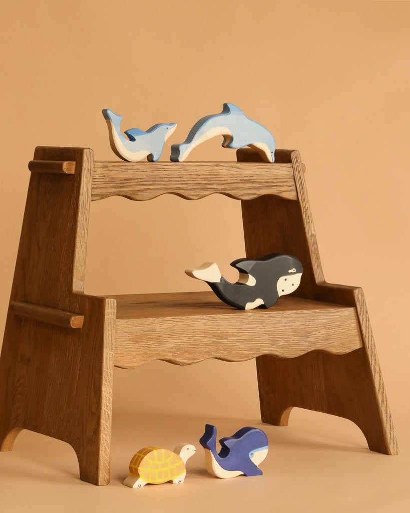The Holztiger Handmade Wooden Ocean Animals Set—including whales, a dolphin, a seal, a turtle, and a penguin—features beautifully painted wooden figures displayed on a step stool against a beige background.