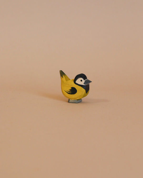 The Ostheimer Goldfinch is a small, handcrafted wooden bird figurine made from sustainably sourced materials. It is painted in yellow, black, and grey tones and features a black head with white cheeks and a yellow body, closely resembling a great tit. The bird is shown against a neutral light beige background.