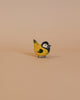 The Ostheimer Goldfinch is a small, handcrafted wooden bird figurine made from sustainably sourced materials. It is painted in yellow, black, and grey tones and features a black head with white cheeks and a yellow body, closely resembling a great tit. The bird is shown against a neutral light beige background.