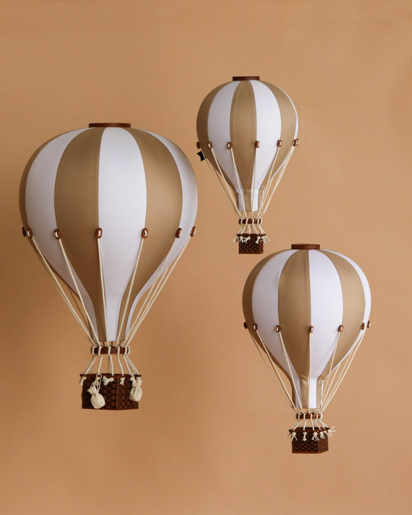 Three decorative handmade Hot Air Balloon - Gold ornaments, featuring alternating white and beige panels with small brown baskets, are suspended against a solid beige background—perfect for charming children’s room decoration.