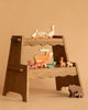 A wooden shelf displays the Holztiger Handmade Wooden Woodland Animals Set—including a goose, deer, squirrel, rabbit, and bear—against a neutral beige background, perfect for open-ended play.