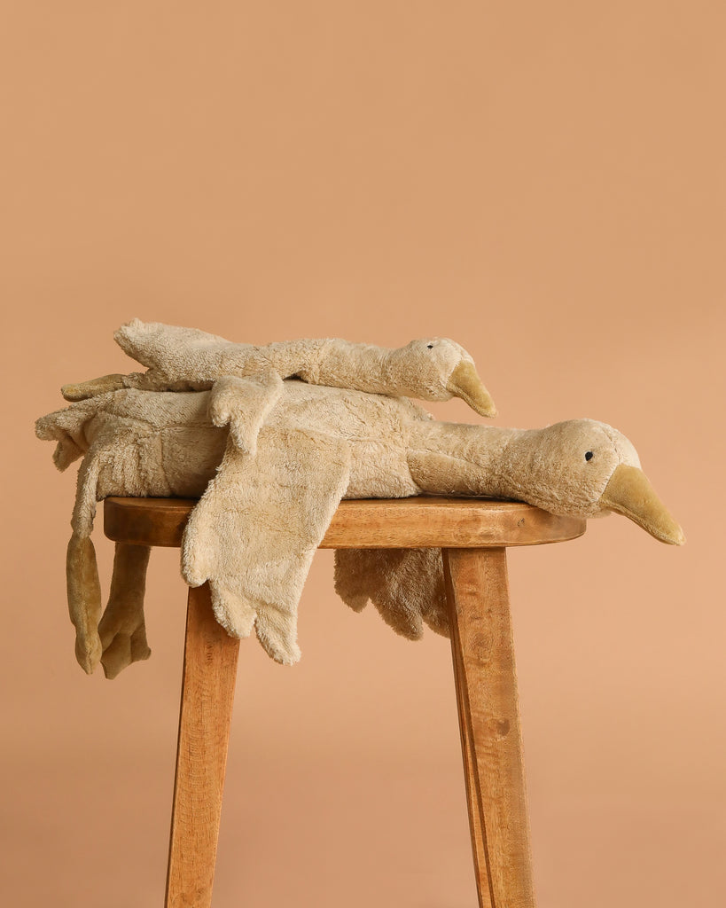 Two Senger Naturwelt Cuddly Animal Linen Geese, made from organic materials, are stacked atop each other on a wooden stool set against a plain beige background.