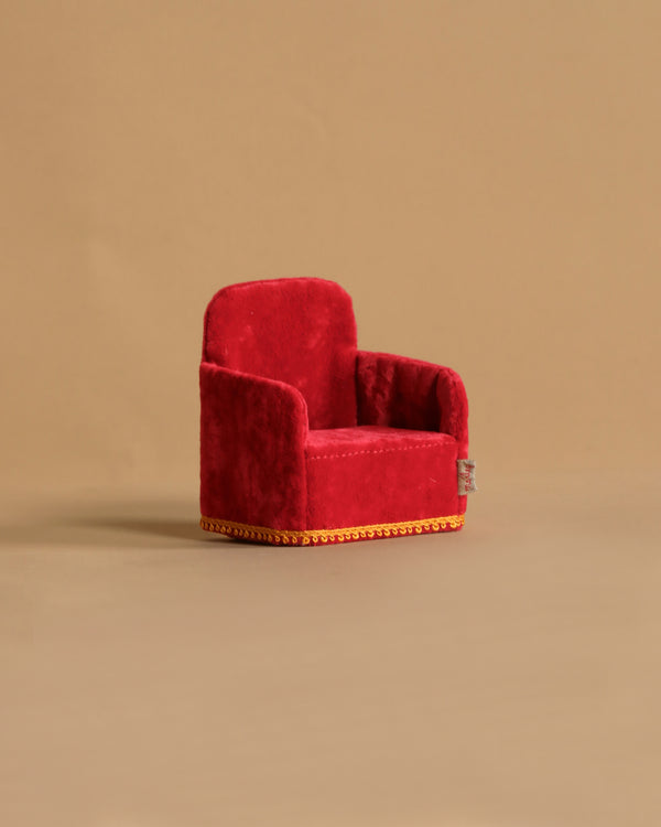 The Maileg Soft Chair - Red, featuring gold trim and a lush texture, rests against a plain beige background. Perfect for Mouse hole Farmhouse settings, it resembles a miniature armchair welcoming your Maileg friends with its vibrant color.