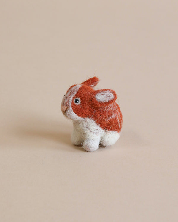 A hand felted bunny with brown and white coloring, positioned against a plain beige background. The bunny is small and detailed, showing a fluffy texture made from wool fibers.