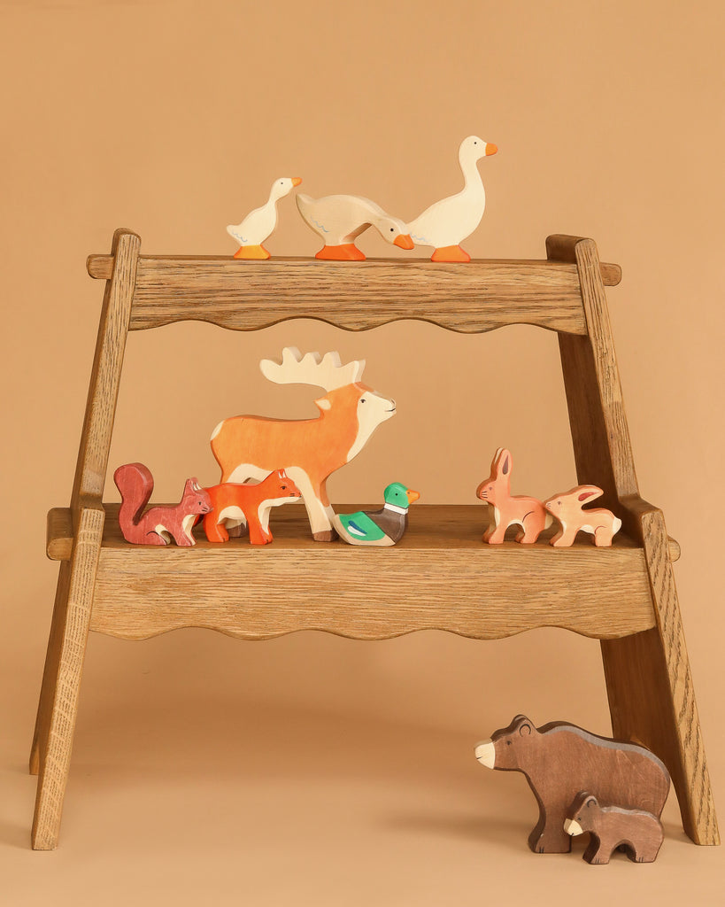 The Holztiger Handmade Wooden Woodland Animals Set features ducks, a deer, squirrels, rabbits, and two bears displayed on a two-tiered wooden stand, perfect for open ended play against a beige background.