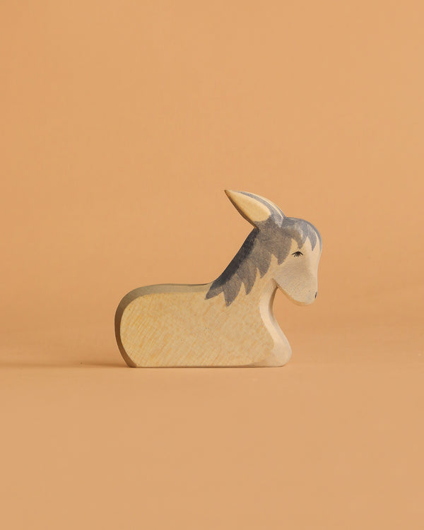 The Ostheimer Donkey - Resting is a small, handcrafted wooden figurine made from sustainable materials and features painted details. It is shown on a light orange surface with a matching orange background.