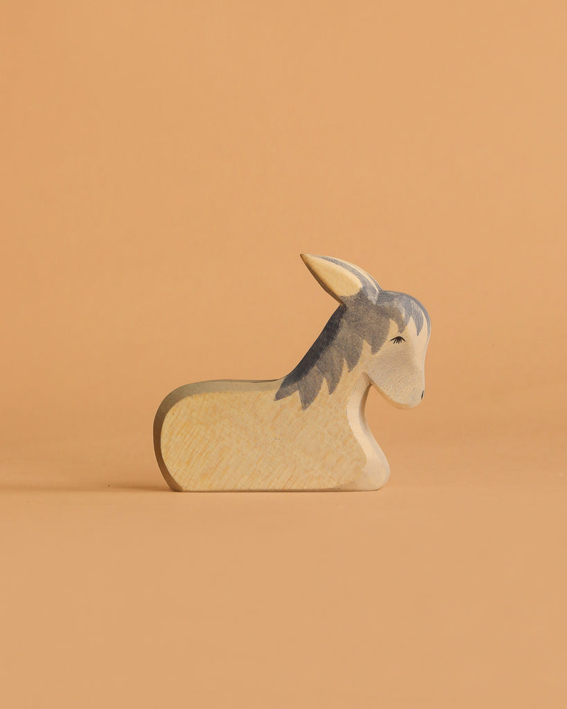 The Ostheimer Donkey - Resting is a small, handcrafted wooden figurine made from sustainable materials and features painted details. It is shown on a light orange surface with a matching orange background.