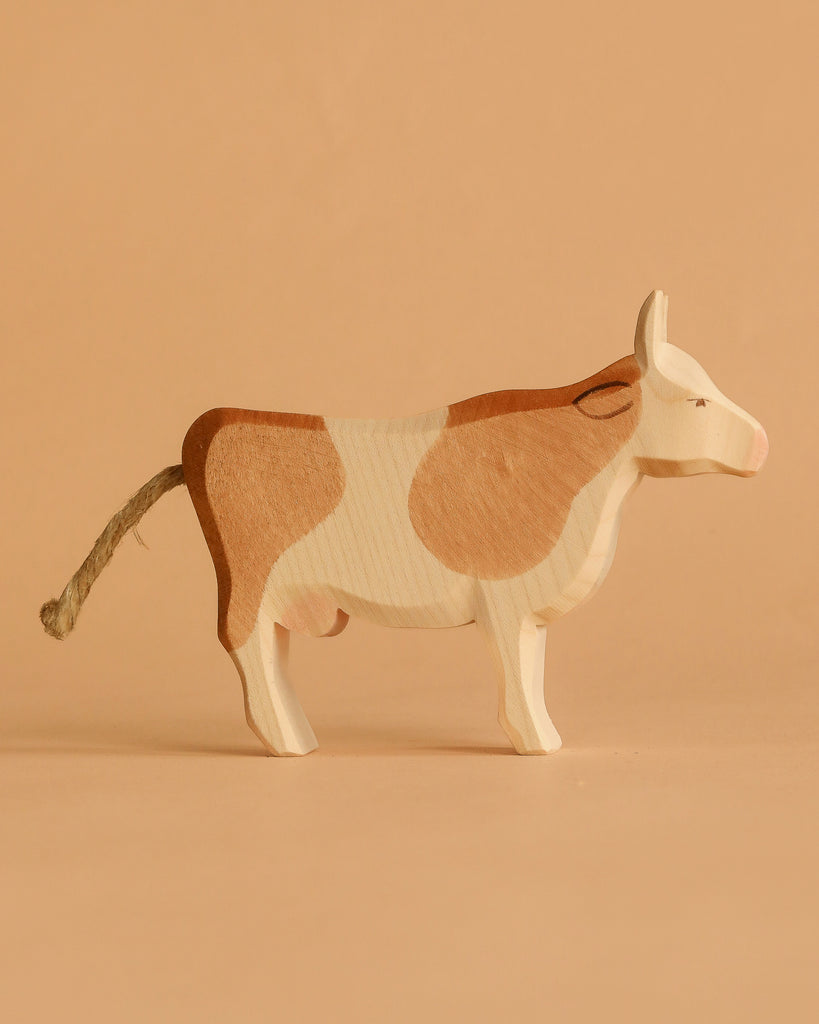 A figurine from the Ostheimer Handmade Wooden Farm Animals Set stands on a beige surface. Crafted from sustainably sourced wood, it features light brown and white patches, a rope tail, and a simple, handcrafted design inspired by classic toys.