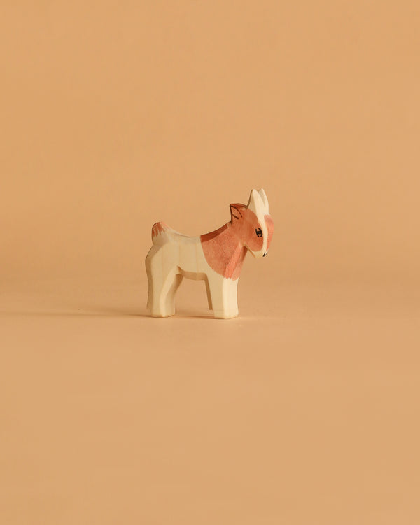 The Ostheimer Goat Kid - Standing is a small, handcrafted wooden figurine painted light brown and white, displayed on a beige surface—ideal for imaginative play or as a charming addition to your wooden toy collection.