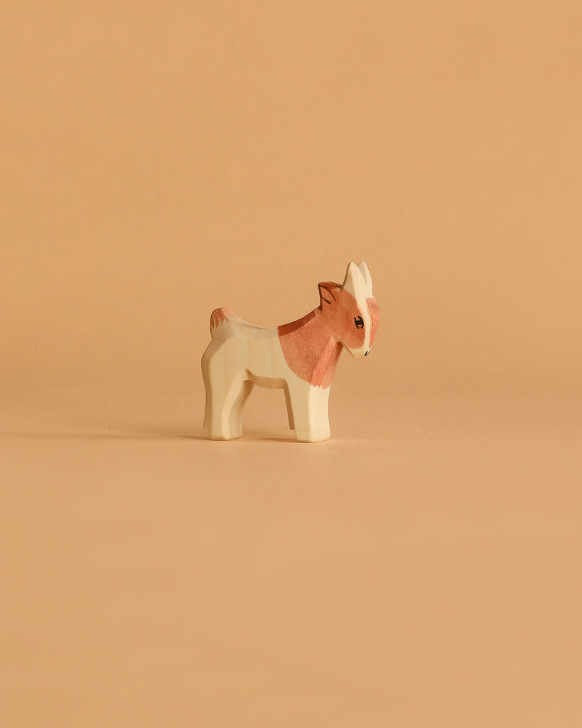A small, handcrafted goat from the Ostheimer Handmade Wooden Farm Animals Set, featuring light brown and white hues, stands on a beige surface—showcasing charming toys made from sustainably sourced wood.