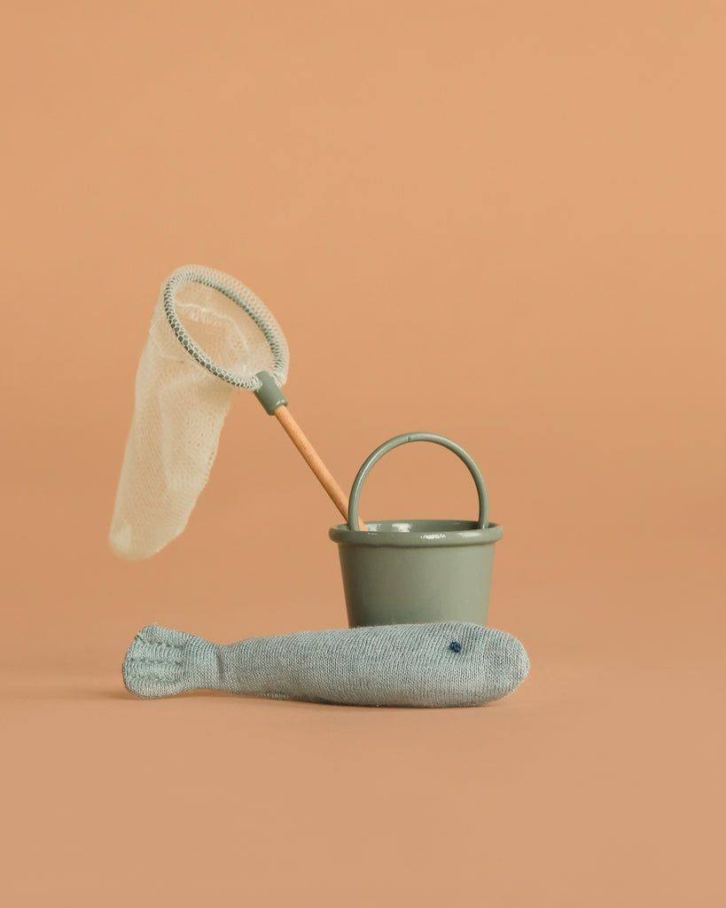 The Maileg Little Brother Gone Fishing Set—featuring a green bucket, wooden-handled fishing net, and soft toy fish—is arranged against a peach background, ready as charming beach house toys for your mouse family.