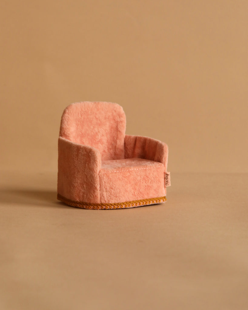 The Maileg Soft Chair - Rose is a small pink plush armchair, perfect for Maileg friends. It stands out charmingly against a plain beige background with its cozy texture, rounded back, and trim-edged base, blending farmhouse charm and playful elegance beautifully.