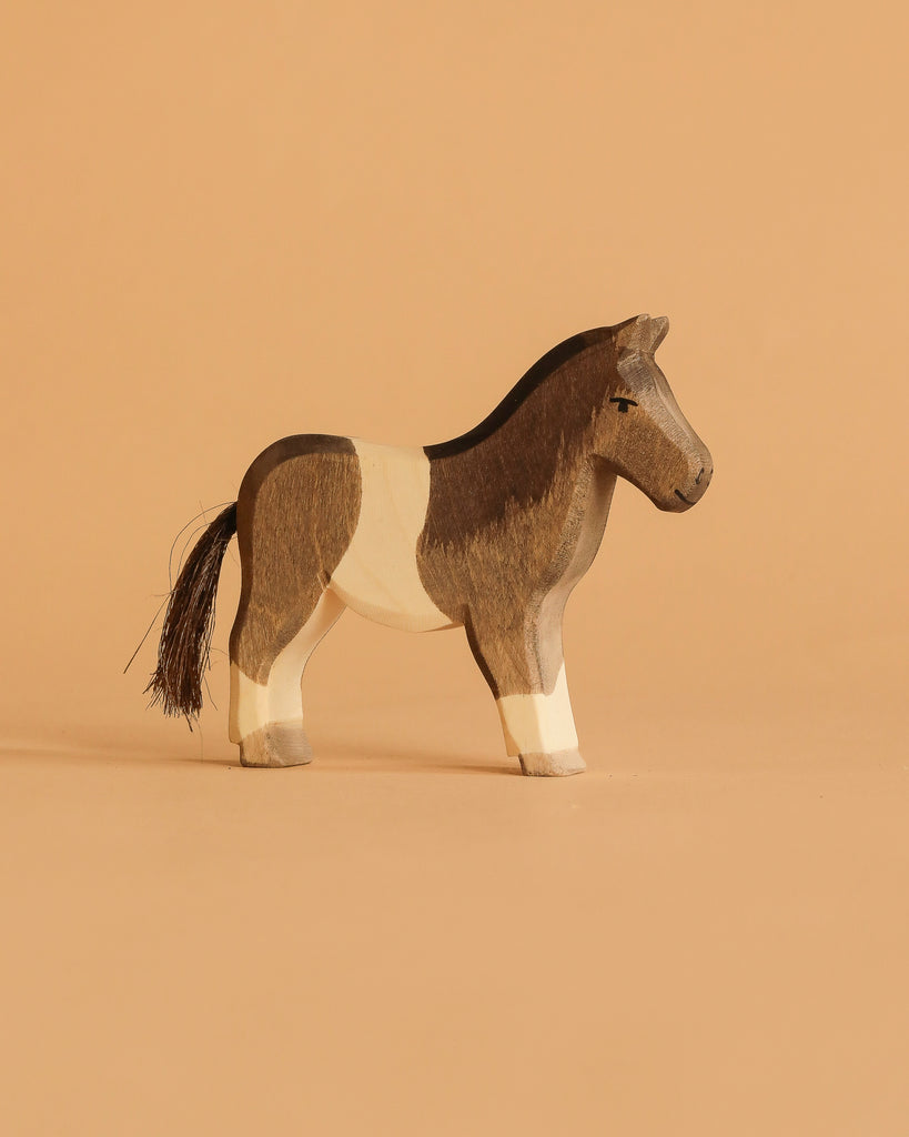 A small, hand-carved horse from the Ostheimer Handmade Wooden Farm Animals Set features brown and cream patches with a black mane and tail, displayed on a plain light brown background.