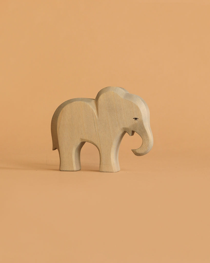 A small, light-toned elephant from the Ostheimer Handmade Wooden Safari Animals Set stands on a matching surface against a plain background, showcasing the charm of handcrafted toys.