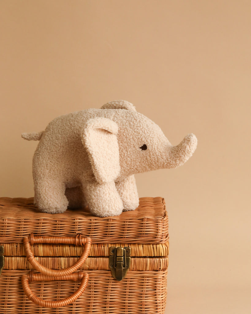 A small, plush Elephant Teddy Stuffed Animal stands on top of a closed wicker basket with a beige background. This soft, snuggly elephant is light grey and features large floppy ears and a short trunk. The wicker basket, equipped with a latch and handle on the front, also has a cozy cotton lining inside.