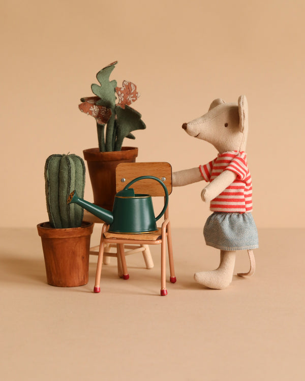 A Maileg Little Green Thumb Garden Set features a mouse in a red striped shirt and blue shorts standing by a flower pot and a green watering can on a school chair, all set against a beige background.