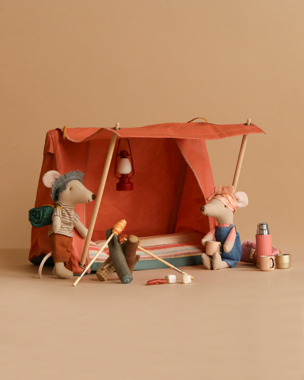 Two toy mice in camping attire are using the Maileg Camping Set, which includes a tent, bonfire, thermos, cups, and lantern. One mouse roasts a marshmallow over the fire while holding a backpack; the other sits nearby with their gear.