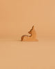 A small, handcrafted wooden deer from the Ostheimer Handmade Wooden Woodland Animals Set, painted with white spots and gray stripes, sits upright on a beige surface against a plain beige background.