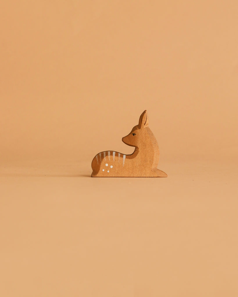 A small, handcrafted wooden deer from the Ostheimer Handmade Wooden Woodland Animals Set, painted with white spots and gray stripes, sits upright on a beige surface against a plain beige background.