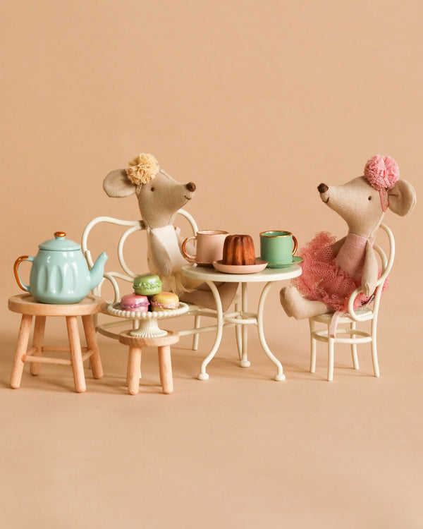 Two Maileg Ballerina Mice in dresses sit at a small table having tea with the Maileg Afternoon Tea Set, which includes cups, cakes, and macarons. The background is a plain beige.