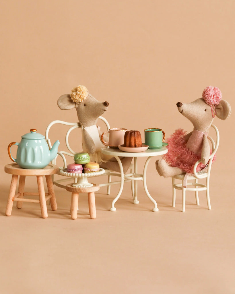 Two Maileg Ballerina Mice in dresses sit at a small table having tea with the Maileg Afternoon Tea Set, which includes cups, cakes, and macarons. The background is a plain beige.