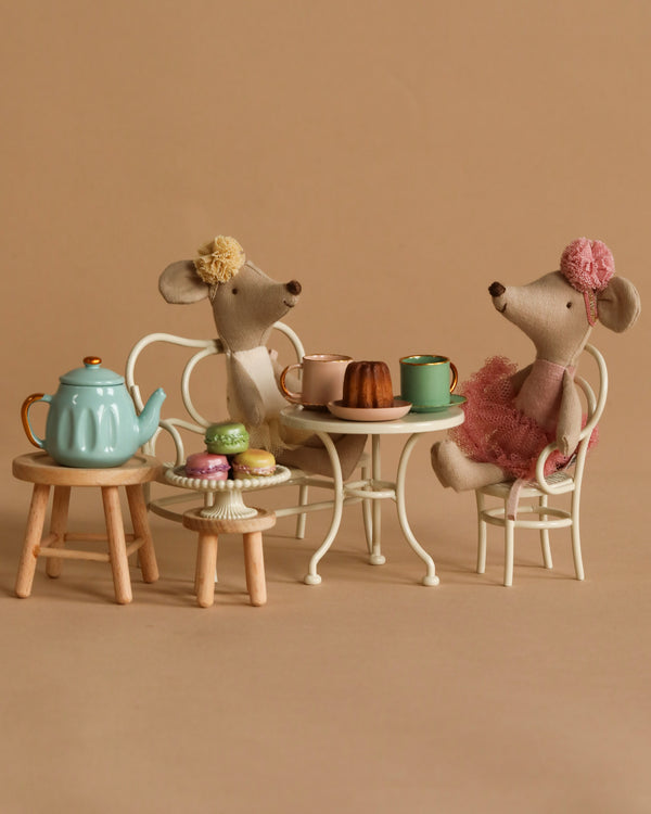 Two Maileg Ballerina Mice, with one in a yellow headband and the other in a pink hat and dress, sit at a small white table set for tea. A teal teapot, mug, cake, and macarons from the Maileg Afternoon Tea Set are on stools beside them against a beige background.