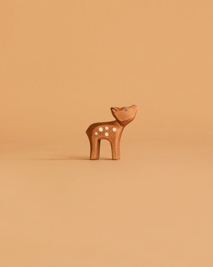 A small, brown deer figurine with white spots stands on a plain, light brown background, reminiscent of the Ostheimer Handmade Wooden Woodland Set.