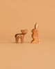 Two small figures from the Ostheimer Handmade Wooden Woodland Set—a deer with white spots and a rabbit—stand facing each other on a plain light brown background.