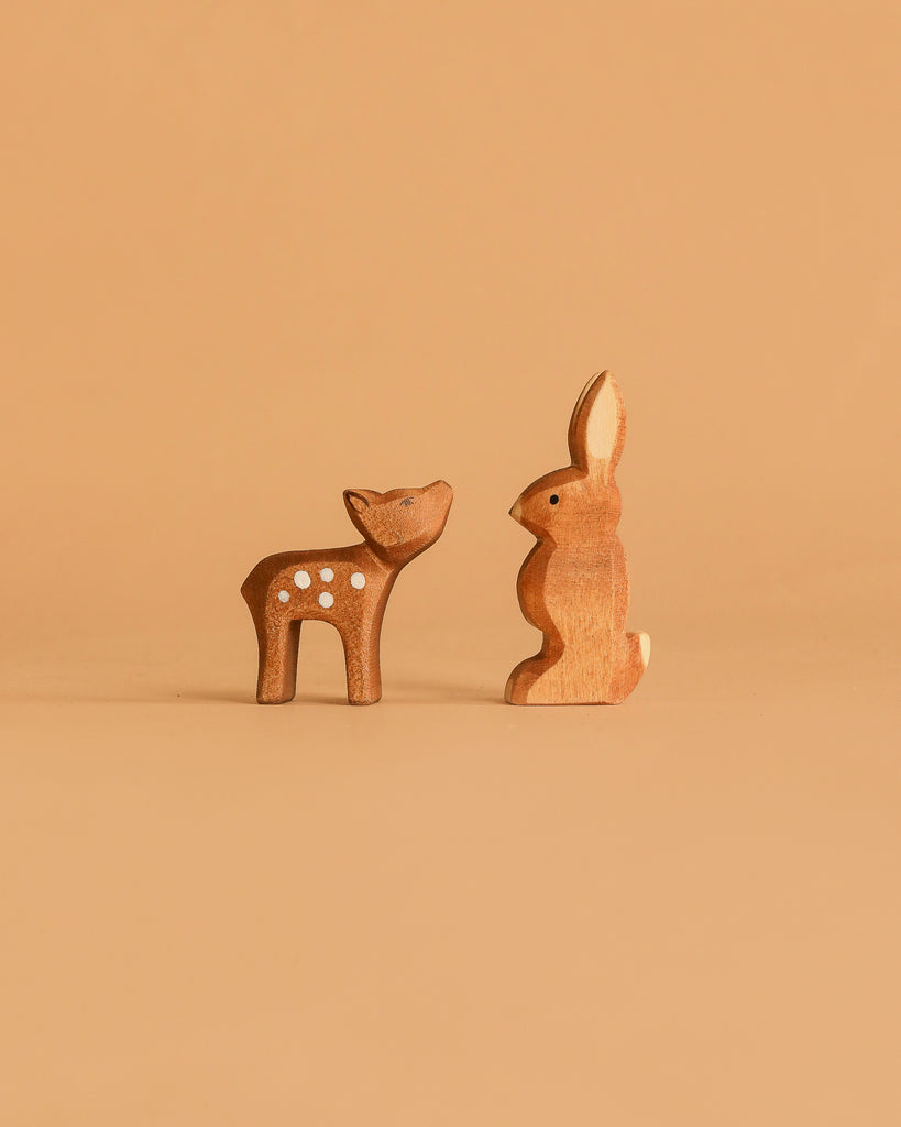Two small figures from the Ostheimer Handmade Wooden Woodland Set—a deer with white spots and a rabbit—stand facing each other on a plain light brown background.