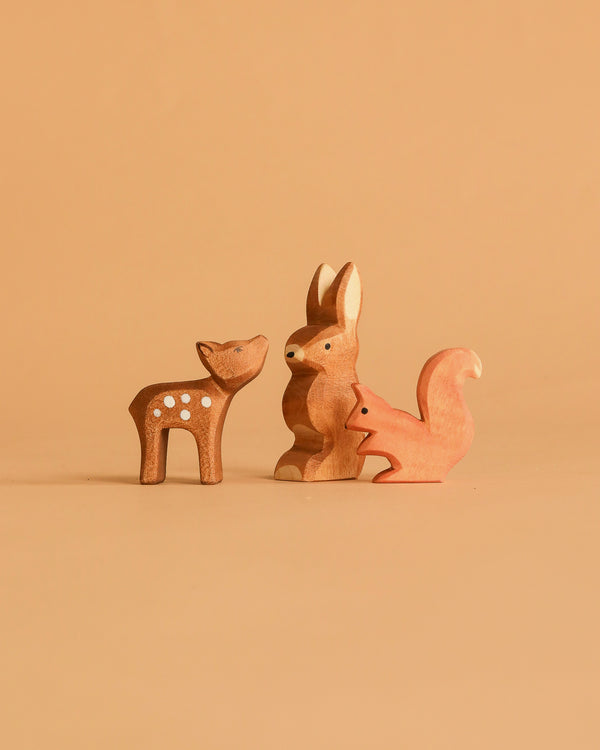 The Ostheimer Handmade Wooden Woodland Set features three handcrafted wooden toys—a deer with white spots, a rabbit in the center, and a squirrel on the right—displayed on a plain, light brown background.