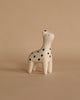 A small, white ceramic figurine of a polka-dotted animal stands on a beige surface. The Handmade Tiny Wooden Safari Animals - Giraffe features black spots and a minimalist facial expression with closed eyes. Reminiscent of handcrafted Albizia wood toys, the background is simple and matches the beige tone of the surface.