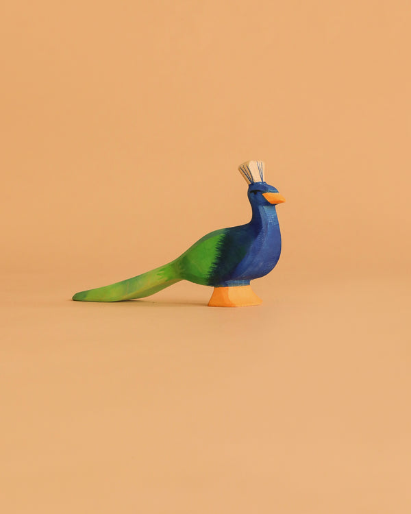 The Ostheimer Peacock, a handcrafted wooden figurine painted in blue, green, and orange, stands on a simple peach background—evoking the classic German-made Ostheimer toy style.