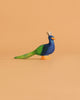 The Ostheimer Peacock, a handcrafted wooden figurine painted in blue, green, and orange, stands on a simple peach background—evoking the classic German-made Ostheimer toy style.