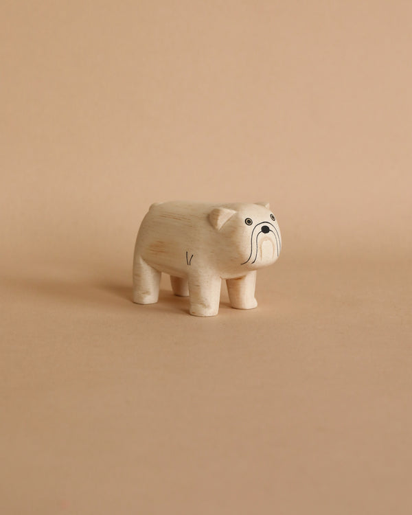 A small, hand-carved wooden bulldog figurine with visible wood grain, featuring large cartoon-style eyes and a curved trunk, stands against a plain beige background.