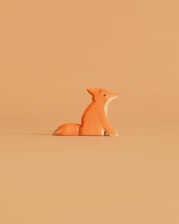 The Ostheimer Small Fox - Sitting is a minimalist, handcrafted wooden figurine painted orange with white accents, displayed on a simple beige surface against a matching beige background.