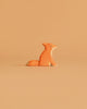 The Ostheimer Small Fox - Sitting is a minimalist, handcrafted wooden figurine painted orange with white accents, displayed on a simple beige surface against a matching beige background.