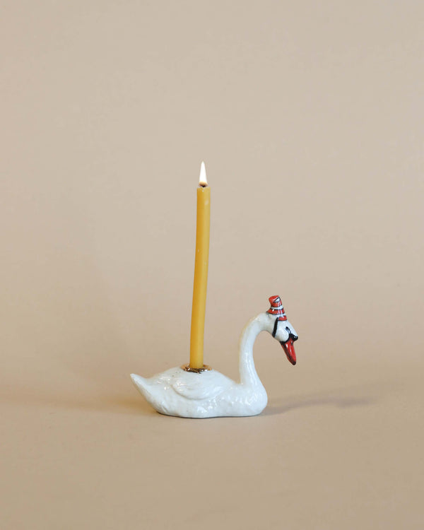 A white hand-painted porcelain Swan Cake Topper with a yellow candle lit, set against a soft beige background. The swan has a small red bow around its neck.
