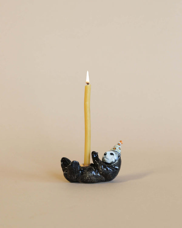 A lit yellow candle is held upright by a small hand-painted porcelain Sea Otter Cake Topper, set against a plain beige background.