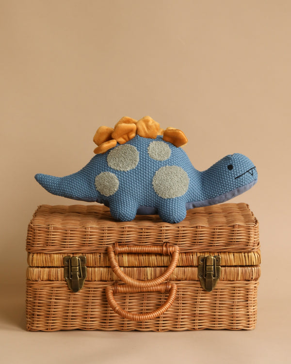 A Stegosaurus Dinosaur Stuffed Animal with blue plush fabric, yellow bumps on its back, and gray spots stands on a closed wicker basket that features a handle and two latches. Suitable for ages 0+, this adorable toy is perfect for little ones. The background is a plain beige color.