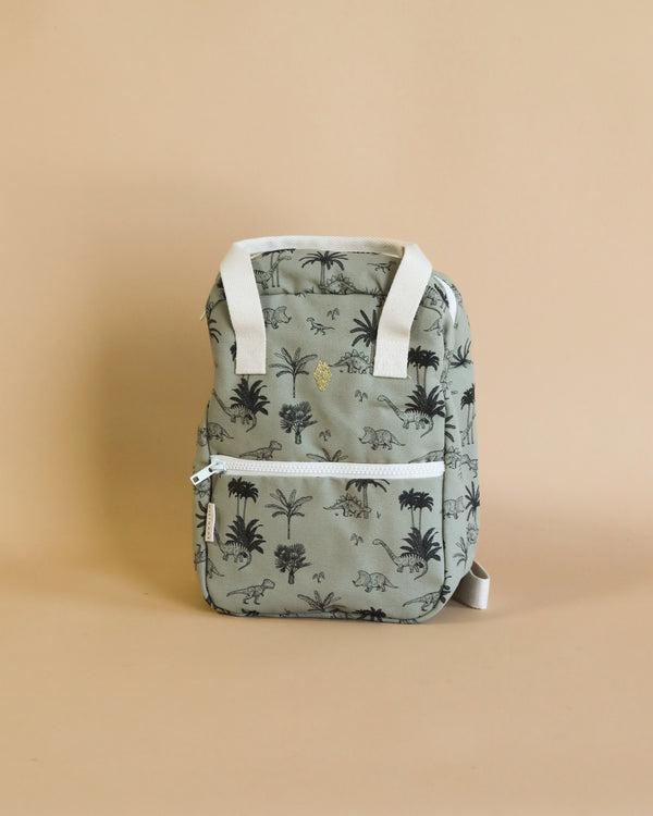 A small toddler Milinane Backpack - Dinosaur with a tropical palm tree and dinosaur print, featuring a top handle and front zip pocket, set against a plain beige background.
