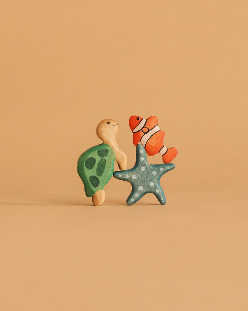 Three toys from the Handmade Holzwald Ocean Animals Set—a green turtle, orange clownfish, and blue starfish with white dots—stand upright on a plain beige background.