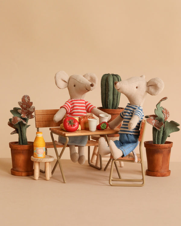 Two Maileg mice from the Picnic in the Garden Set wear red and blue striped shirts and sit at a small table with food, drinks, and potted plants, creating a cozy, playful scene.