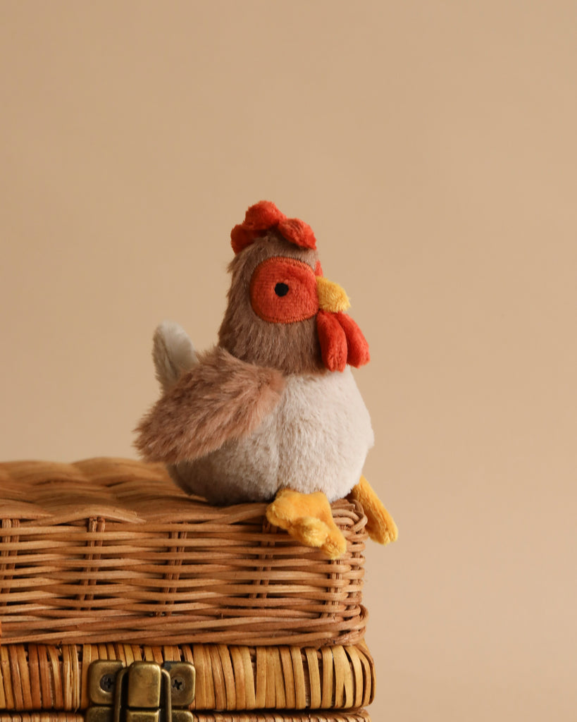 A Bubba Rooster Rattle, a plush toy resembling a chicken, sits atop a closed wicker basket against a plain beige background. The toy, which doubles as a baby rattle, features a red comb and wattle, an orange beak, and detailed feather-like textures in shades of brown and white.