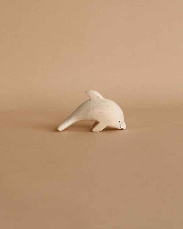 A hand-carved wooden dolphin figurine on a plain beige background, positioned as if swimming to the left, with visible wood grain and a soft matte finish.