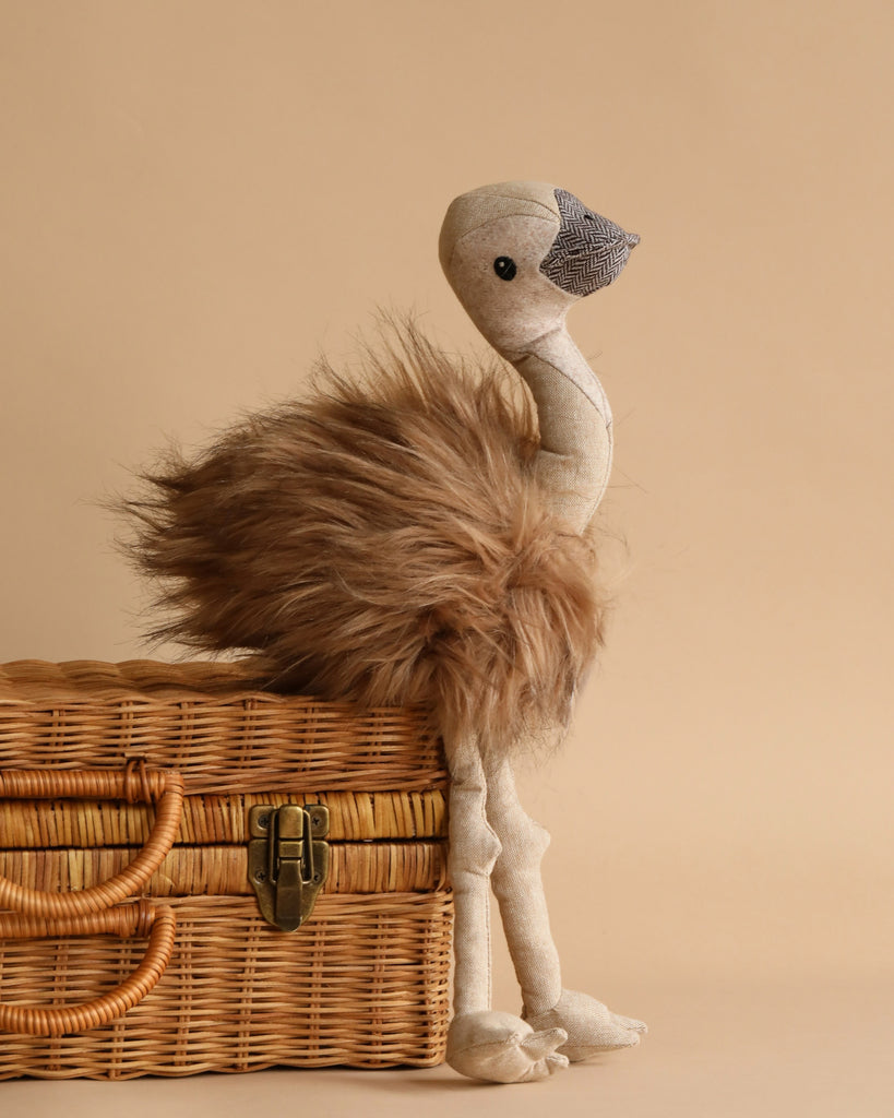 Eddie The Emu Stuffed Animal, a handmade toy featuring a fluffy brown body, long beige legs, and a fabric face with a stitched beak and eyes, stands next to a wicker basket. Suitable for children aged 3 and up, it measures 45cm x 15cm. The background is a plain light beige color.