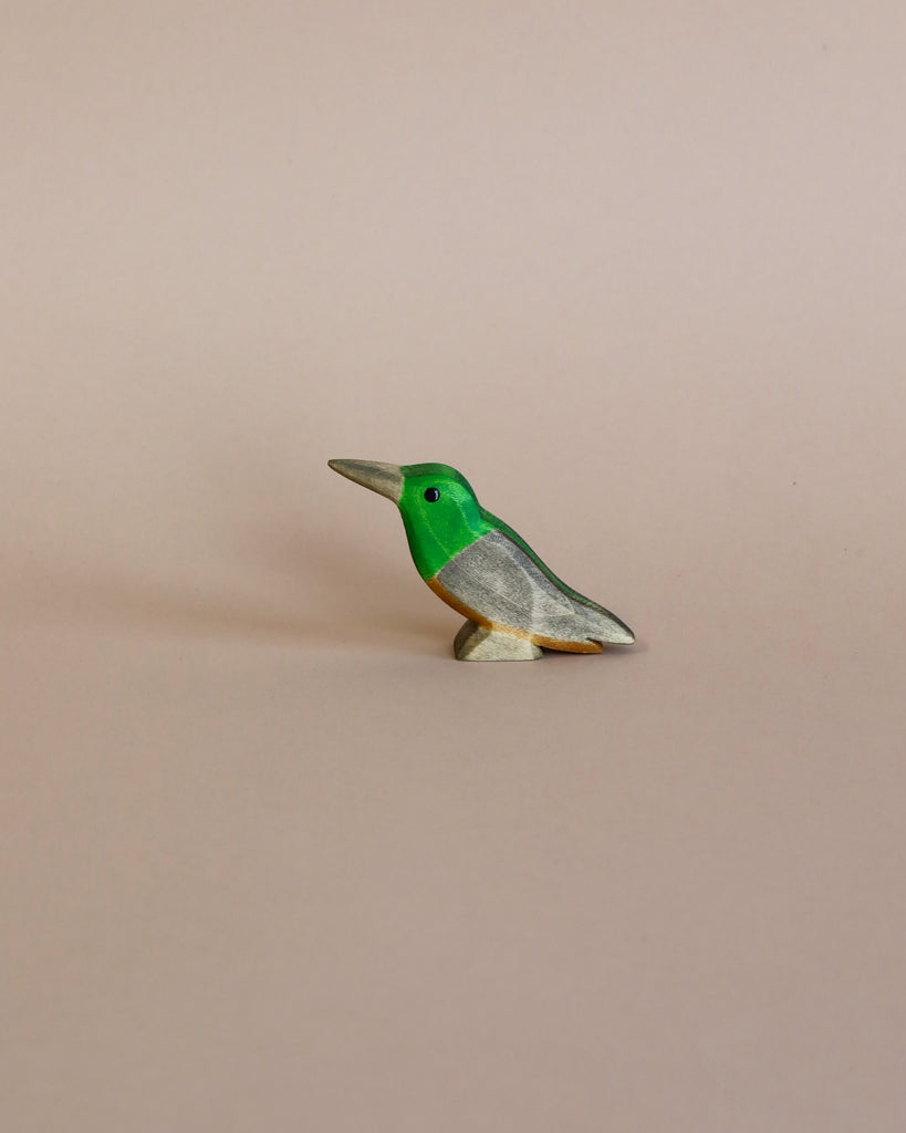 A small Handmade Holzwald Kolibri Bird figurine with a green head and grey body, set against a plain light beige background, epitomizes sustainable wooden toys.