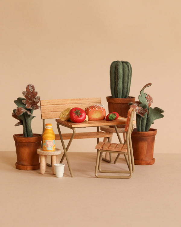 The Maileg Garden Brunch Table Set features a small wooden table and chairs with bread, tomatoes, and a drink bottle, surrounded by potted cacti and plants in mouse size, all arranged against a beige background.