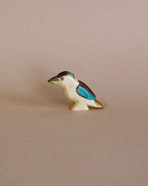 A colorful Handmade Holzwald Kookaburra Bird figurine with teal and white patches stands against a plain beige background. The bird is depicted in a simplistic, high-quality, stylized design.