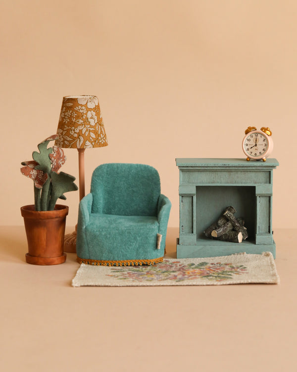 The Maileg Cozy Living Room Set features a teal armchair, floral rug, potted plant, patterned lamp, blue fireplace with logs, and a small alarm clock on the mantel, all set against a beige backdrop.